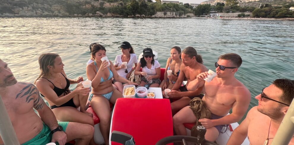 A group of friends enjoying the Devil’s Saddle boat tour, sitting around a small table with snacks and drinks, surrounded by scenic hills and calm waters.