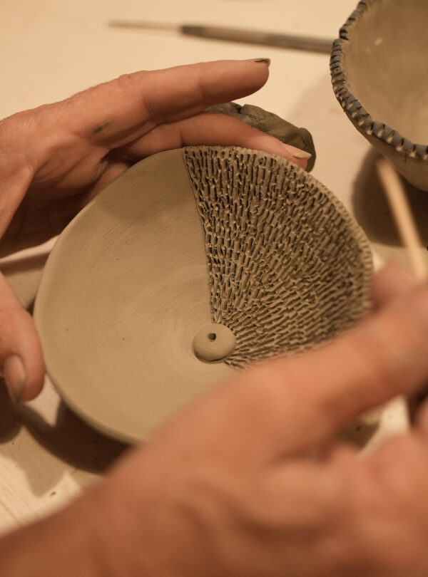 Hands carving textured patterns into a clay bowl using a wooden tool, with another unfinished bowl in the background.
