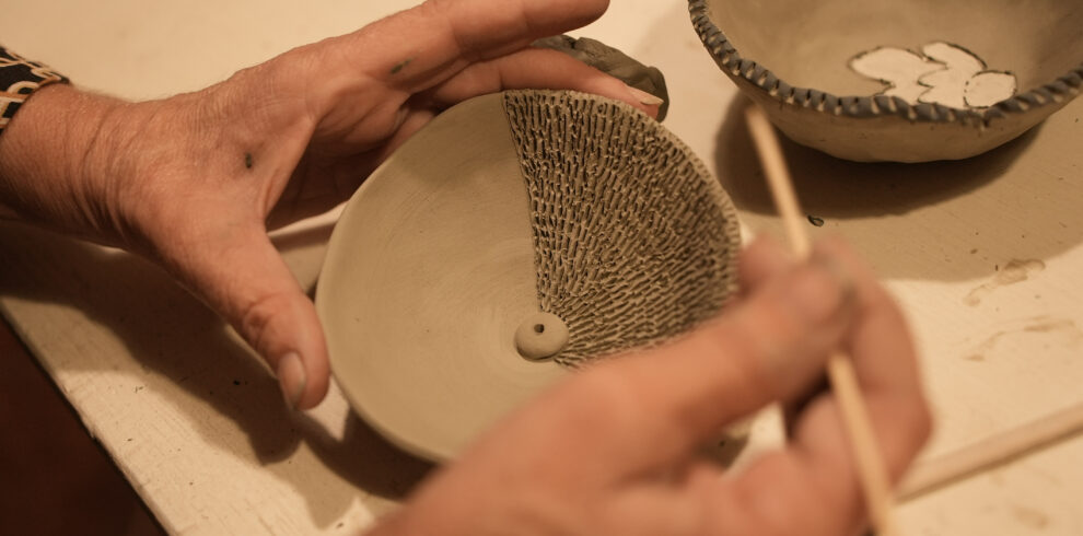Hands carving textured patterns into a clay bowl using a wooden tool, with another unfinished bowl in the background.
