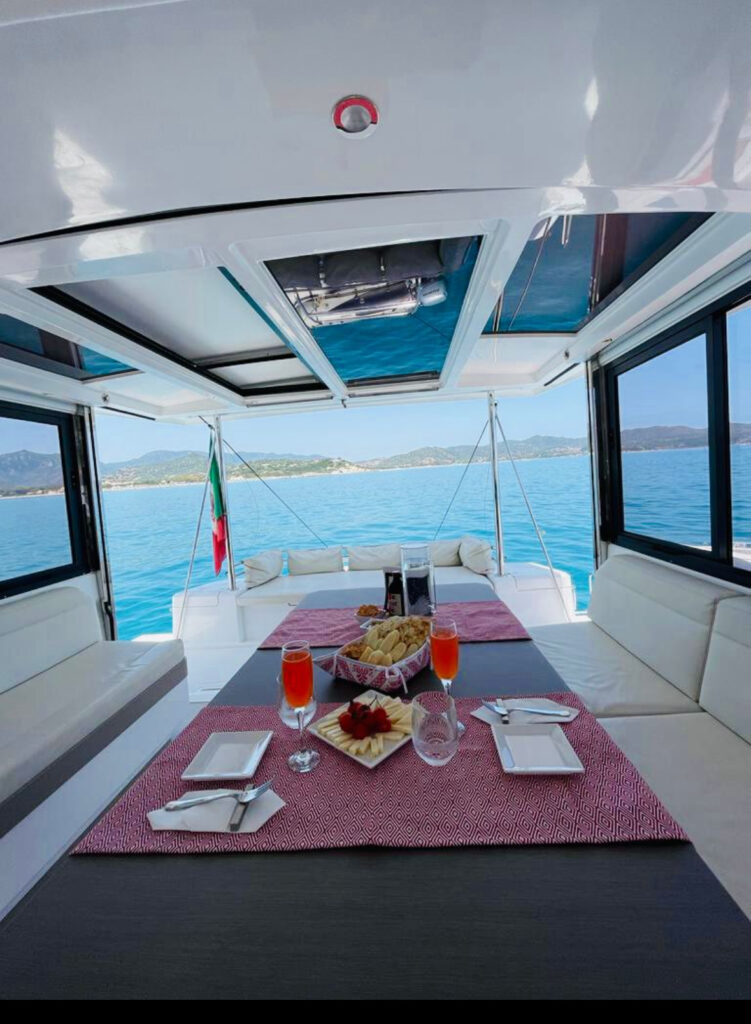 Table set with drinks, bread, and fruit aboard a private catamaran on calm blue waters with a coastal view in the background.